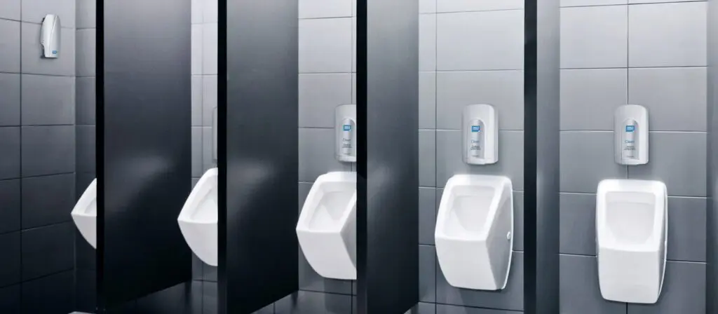 A row of clean, modern urinals in a tiled public restroom, each separated by black partitions. Above each urinal, a wall-mounted soap or sanitiser dispenser is visible, suggesting a focus on hygiene. The restroom has a sleek and minimalistic design.