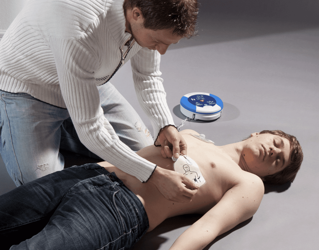 A person applies defibrillator pads to the chest of an unconscious individual lying on the ground. A defibrillator device is placed nearby, and the scene suggests an emergency situation. The rescuer is focused on positioning the pads correctly, preparing to deliver an electric shock.