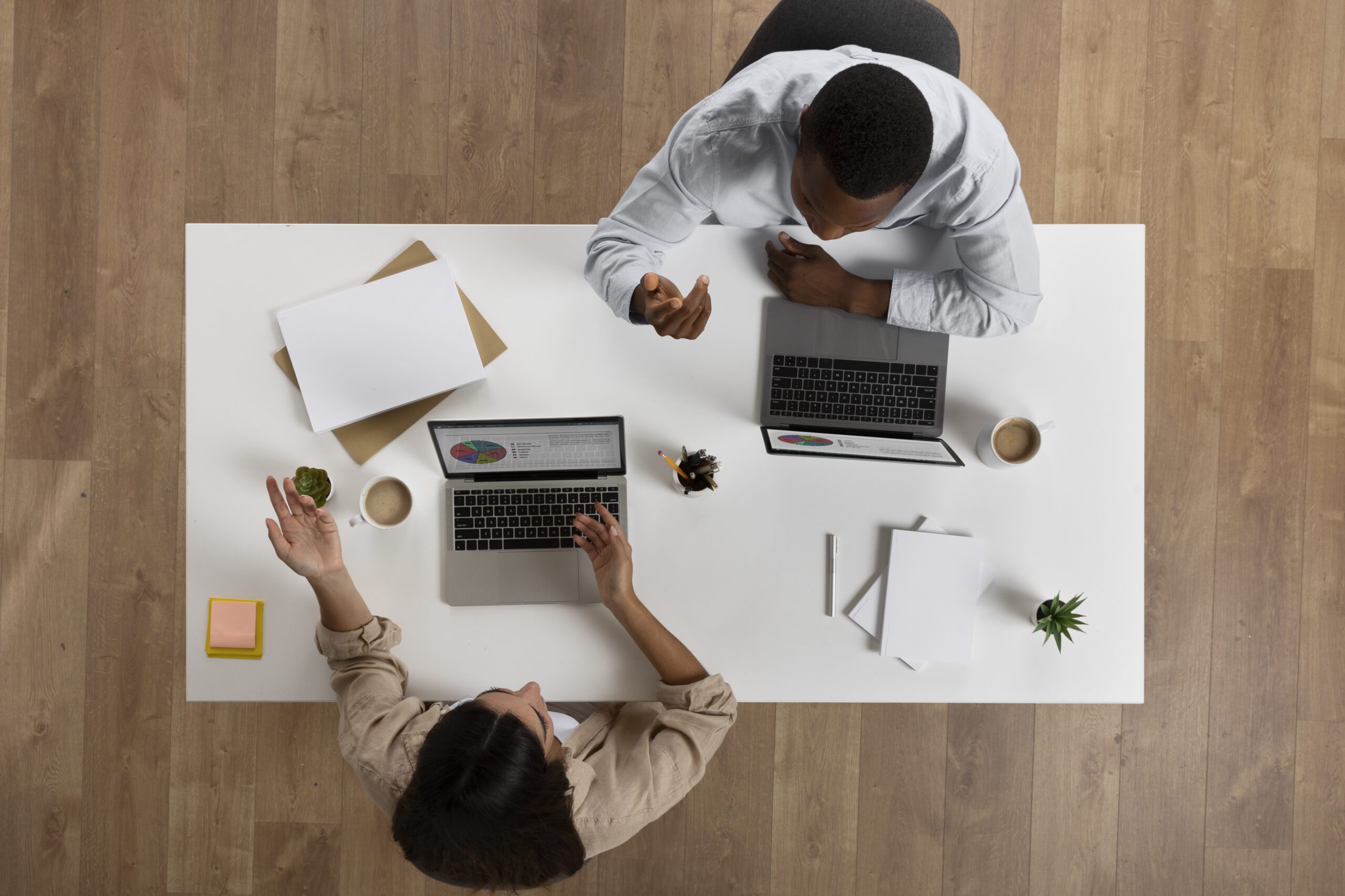 The image shows an overhead view of two individuals sitting at a white desk. Both are working on laptops, with one person pointing towards something on their screen while the other observes or discusses the point. The desk has a few scattered papers, a small plant, and some office supplies, indicating a professional setting. The wooden floor beneath adds warmth to the modern, minimalist office environment.