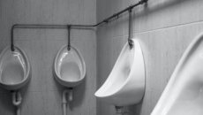 A row of clean, modern urinals in a tiled public restroom, each separated by black partitions. Above each urinal, a wall-mounted soap or sanitiser dispenser is visible, suggesting a focus on hygiene. The restroom has a sleek and minimalistic design.